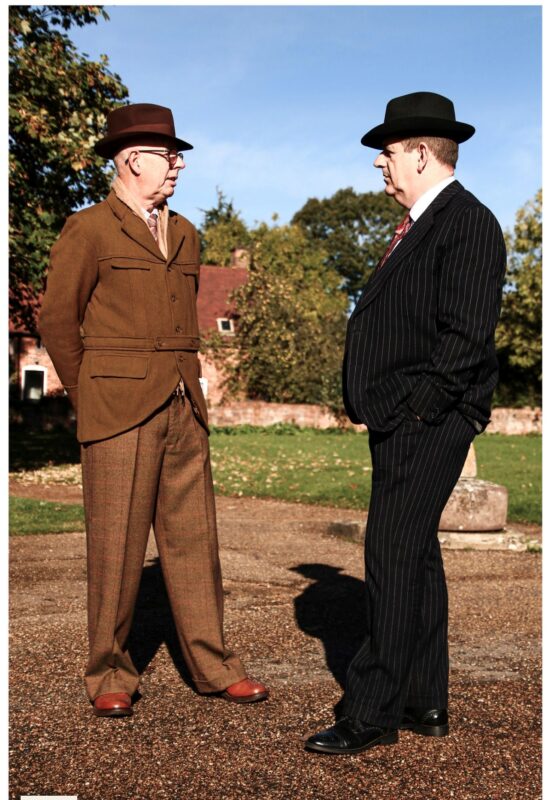 a man dressed in a brown 1940s suit and hat talking to a man dressed in a black 1940s hat and suit