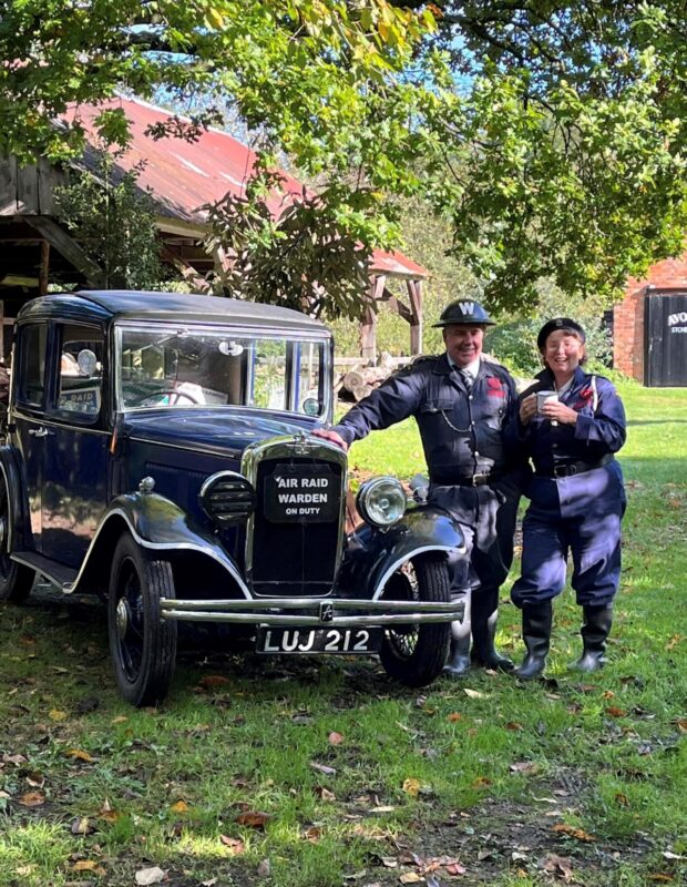 black 1940s car with a a man and women dressed as air raid wardens standing next to it 