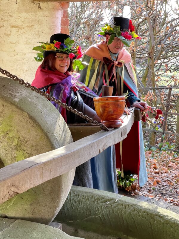 two adults in brightly coloured hats and cloaks pushing the historic jenny ring a cider press. 