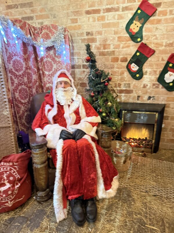 Santa sat in a chair next to fire with stockings on the brick wall behind him