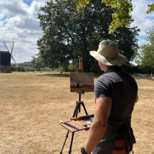 man in foreground at a easel painting a picture of a windmill in the distance in a field