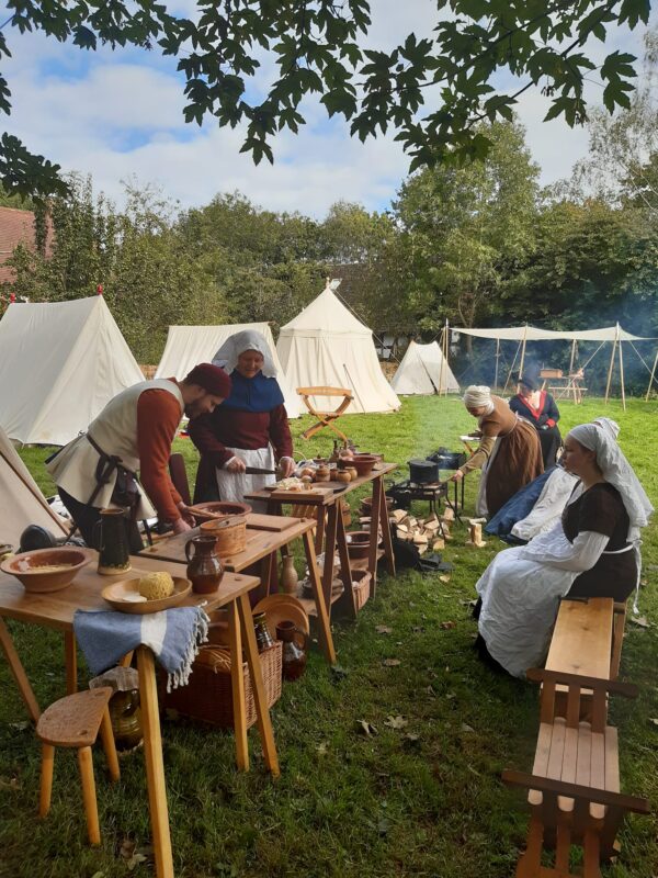 women in medieval clothing preparing food on tables by historical tents on a field.